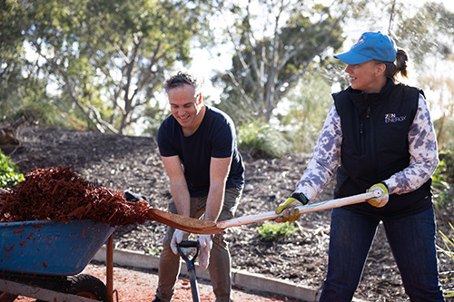 A man and woman smiling at each other while gardening. The woman is wearing a blue heat and a black vest with ZEN Energy's logo. The man is standing next to her wearing a black T-shirt